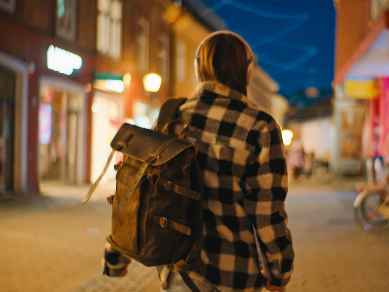 A woman on her own walking away in a lit street in the evening carrying a back pack 